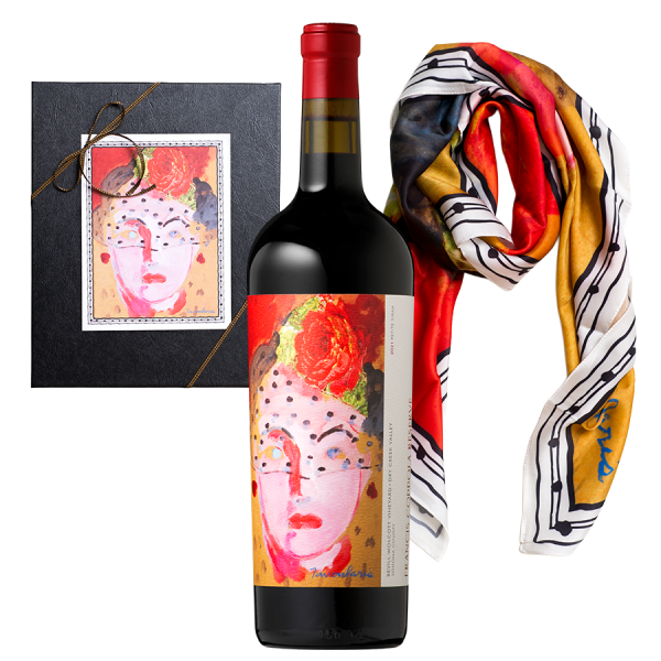 A wine gift set features a bottle with a vibrant, abstract face label, paired with a colorful scarf and a black box. The scene conveys elegance and creativity.