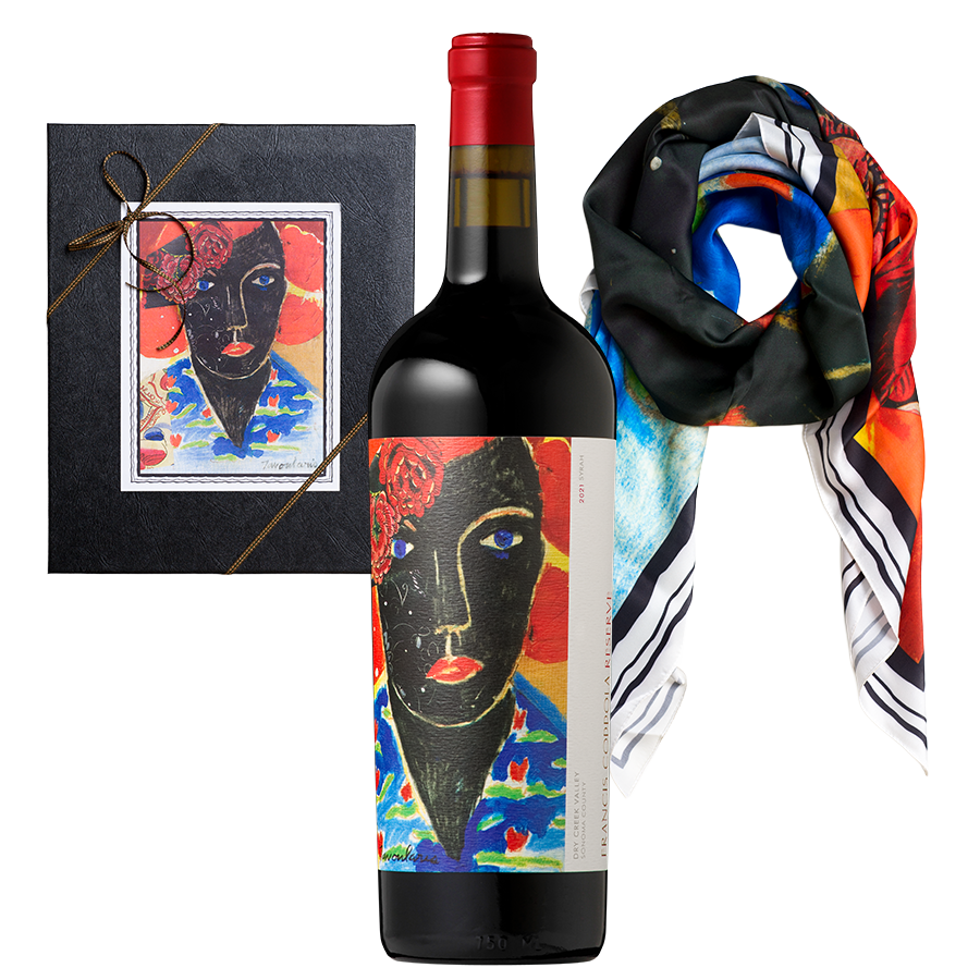 A wine bottle with a bold, colorful abstract face label stands between a black gift box with matching artwork and a vibrant, multi-colored scarf.