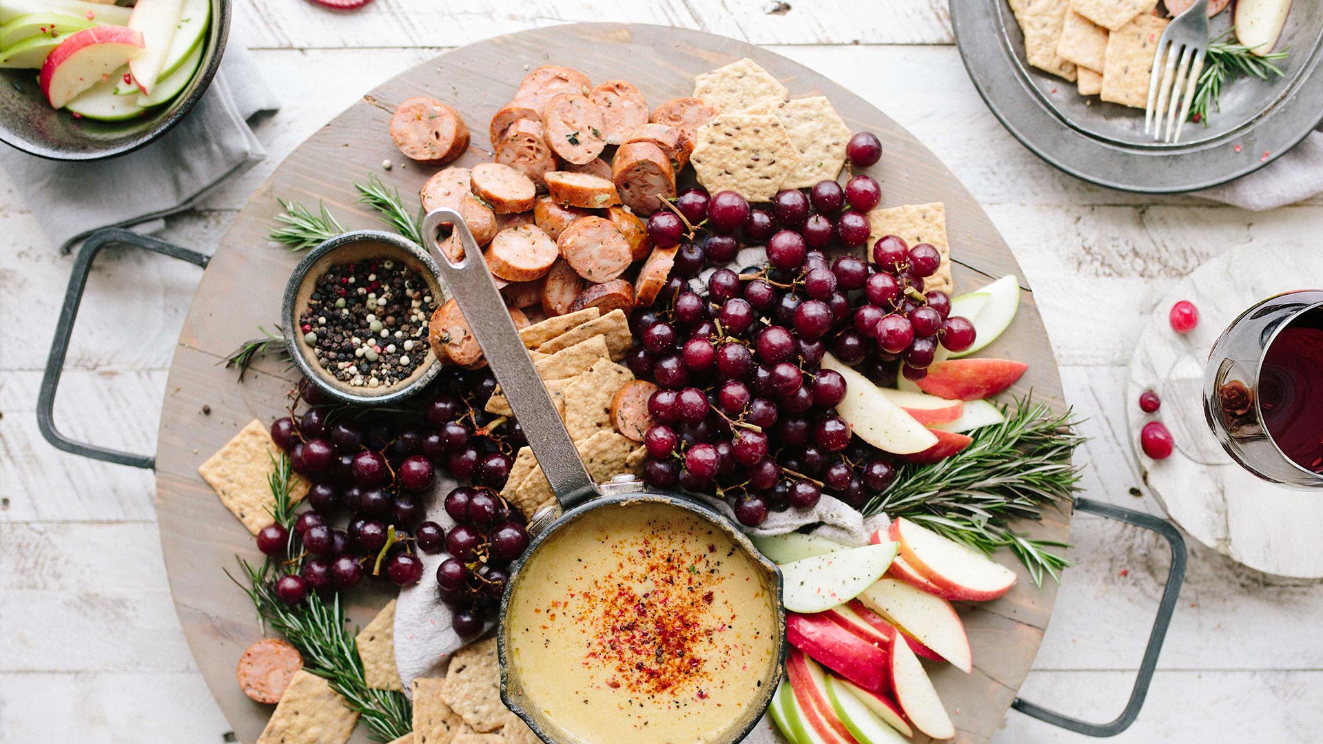 Charcuterie board with sliced sausage, red grapes, apples, crackers, and a skillet of cheese fondue. A glass of red wine is on the side. Inviting and rustic.