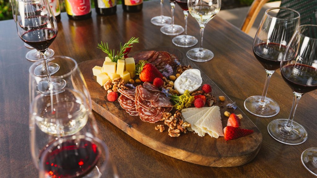 A wooden board with assorted cheeses, sliced meats, strawberries, and nuts is surrounded by glasses of red and white wine on a wooden table.
