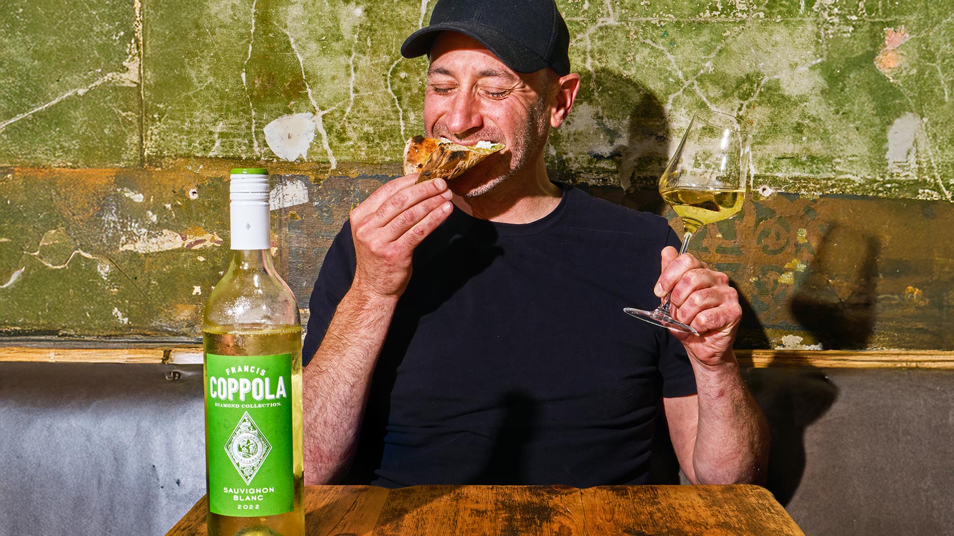 Man enjoying a slice of pizza with eyes closed in delight, holding a glass of white wine. A bottle of wine sits on the rustic wooden table.