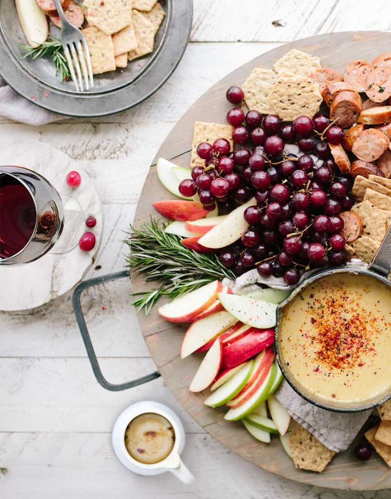 Charcuterie board with sliced sausage, red grapes, apples, crackers, and a skillet of cheese fondue. A glass of red wine is on the side. Inviting and rustic.