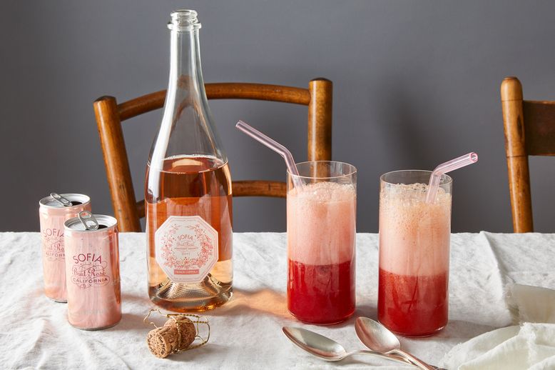 Two tall glasses with pink fizzy drinks, pink straws, and a bottle of wine on a white tablecloth. Two cans and a cork are beside the glasses.