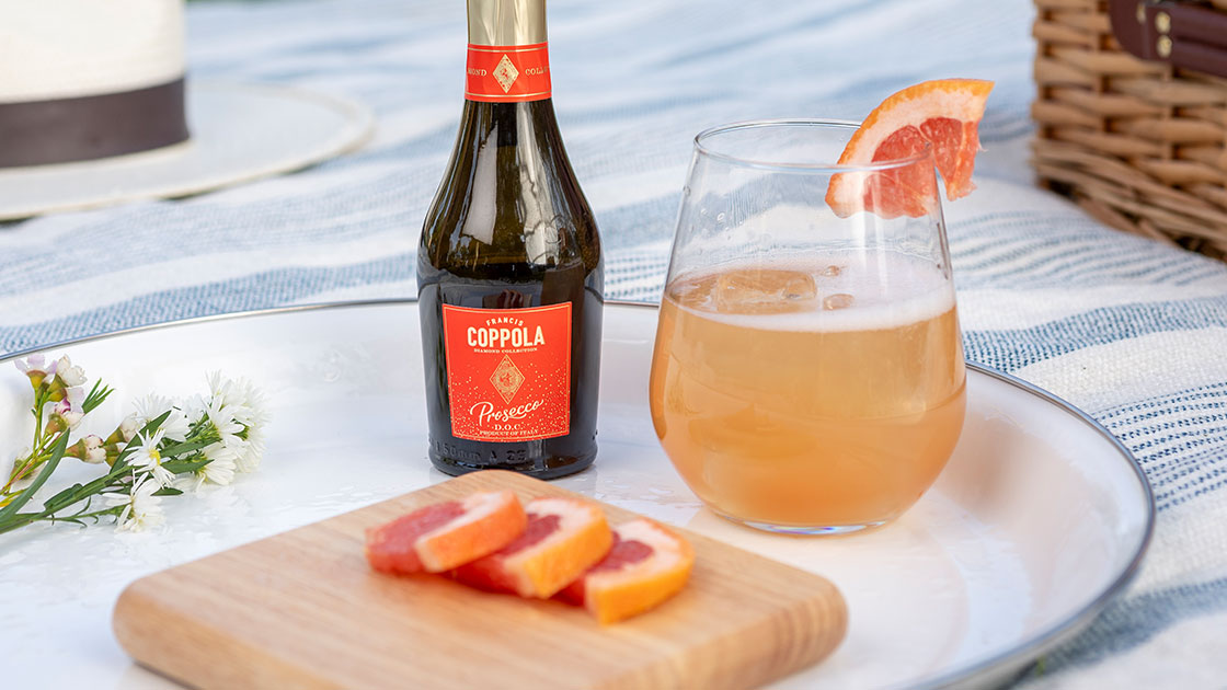 A mini bottle of Coppola Diamond Prosecco pictured with a cocktail glass, slices of grapefruit on a plate in an outdoor picnic setting