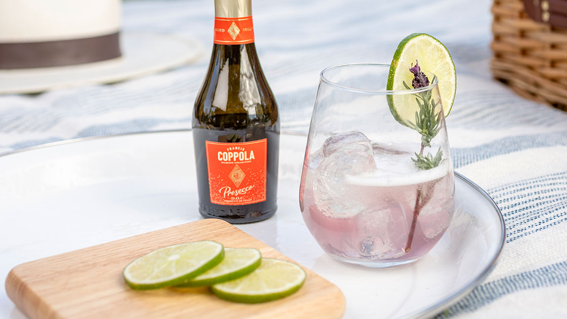 A mini bottle of Coppola Diamond Prosecco pictured with a cocktail glass, slices of limes on a plate in an outdoor picnic setting