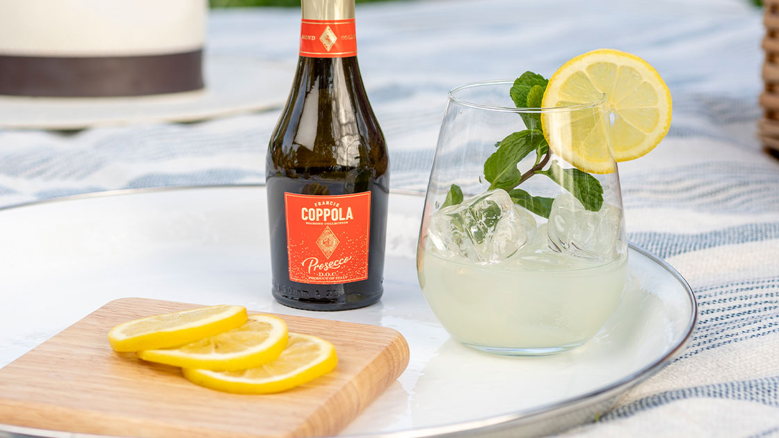 A mini bottle of Coppola Diamond Prosecco pictured with a cocktail glass, slices of lemons on a plate in an outdoor picnic setting